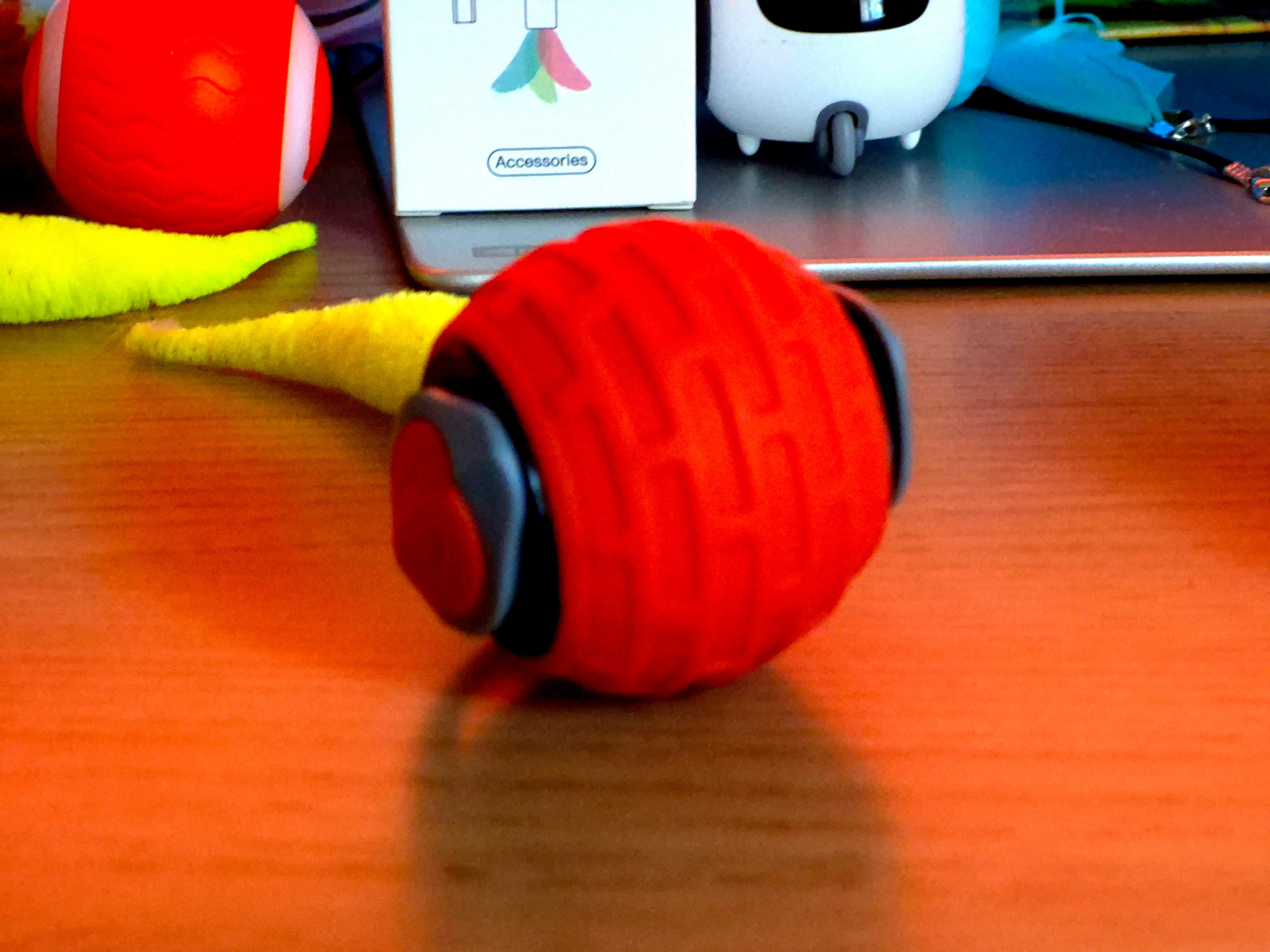 Orange and black textured ball on a wooden surface with a toy and box in the background.